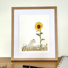 Load image into Gallery viewer, A framed art print featuring a yellow sunflower and a dog in a grassy field, displayed on a wooden desk against a light cream wall.