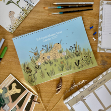 Load image into Gallery viewer, A spiral-bound 2027 calendar featuring an illustration of a soft coated wheaten Terrier in a grassy field, styled on a wooden desk with stationery items.