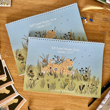 Load image into Gallery viewer, Two spiral-bound 2027 calendars featuring an illustration of a soft-coated wheaten terrier in a grassy field, displayed on a wooden desk with stationery.