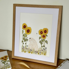 Load image into Gallery viewer, A framed art print featuring a whimsical illustration of a soft coated wheaten terrier amongst yellow sunflowers, displayed on a wooden table against a light wall.