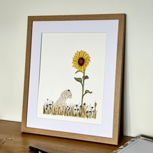 Load image into Gallery viewer, A framed art print featuring a whimsical illustration of a large yellow sunflower and a dog in a grassy field, displayed on a wooden desk against a light wall.
