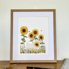 Load image into Gallery viewer, A framed art print featuring a whimsical illustration of yellow sunflowers,  a wheaten terrier, wild flowers and bees, displayed on a wooden desk against a cream wall.
