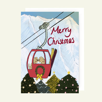 Christmas card with a red gondola lift in a snowy mountain landscape, featuring 'Merry Christmas' text and two Soft Coated Wheaten Terrier Dogs. They are looking down over some decorative Christmas trees.