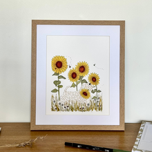 Load image into Gallery viewer, A framed art print featuring a whimsical illustration of a soft coated wheaten terrier among yellow sunflowers, displayed on a wooden tabletop against a light cream wall.
