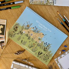 Load image into Gallery viewer, A spiral-bound illustrated calendar lies on a wooden desk, surrounded by stationery items including markers, paintbrushes, a weekly planner, and other art notebooks. The calendar cover shows a soft coated wheaten terrier dog among green grass under a pale blue sky.
