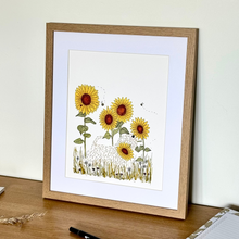 Load image into Gallery viewer, A framed art print featuring an illustration of sunflowers and a soft coated wheaten terrier, displayed on a wooden desk against a light cream wall.
