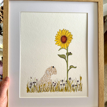 Load image into Gallery viewer, A hand holds a framed art print featuring a whimsical illustration of a dog beside a large yellow sunflower in a grassy field. The print is matted with a white mat board and enclosed in a light oak wood frame.

